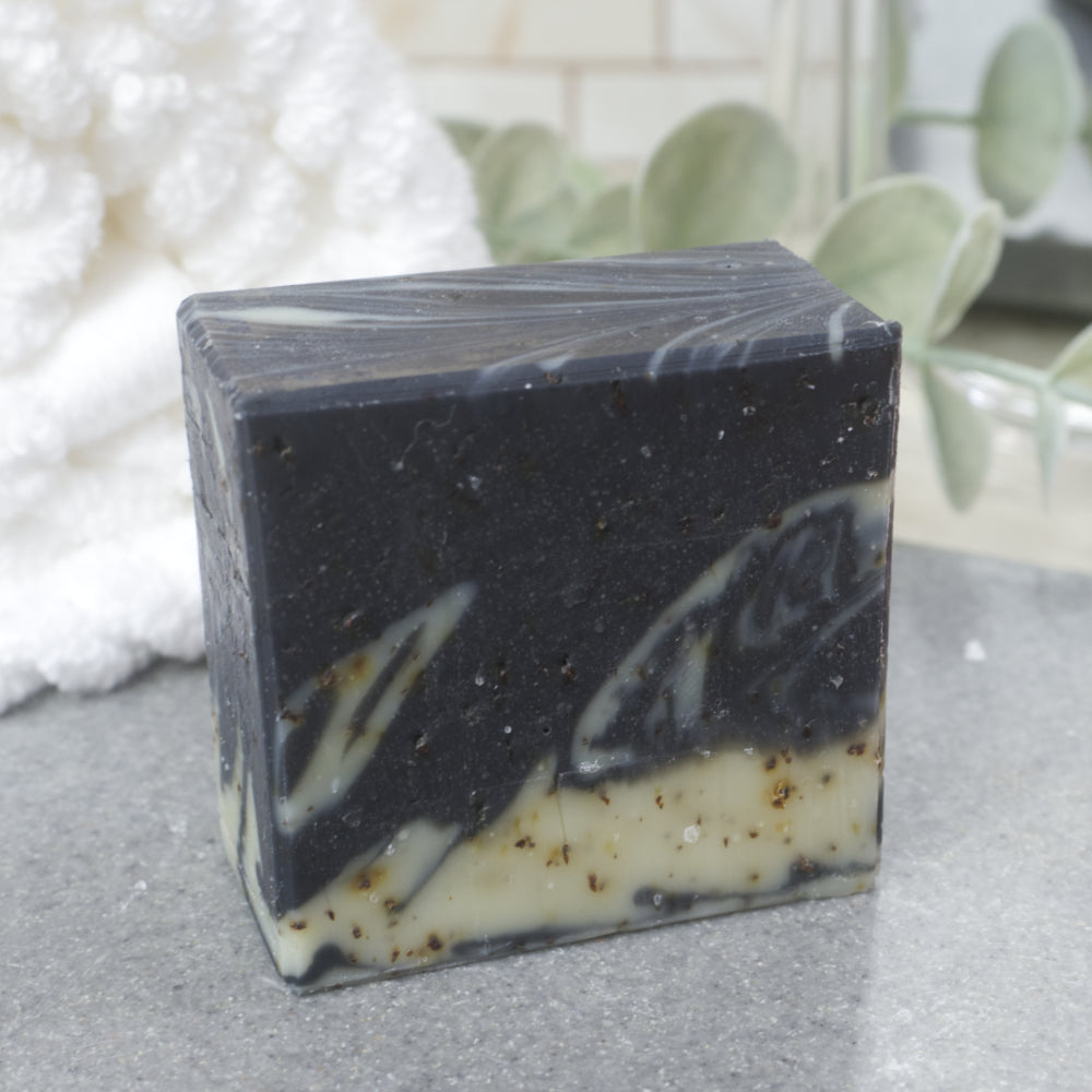A close up image of a single soap bar. It is square in shape with a main colour of black with pale green swirls. It stands on a grey stone tray surrounded by a white towel and greenery.