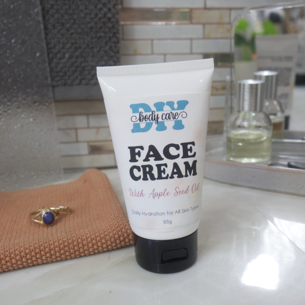 A white squeeze tube of DIY body care Face Cream with Apple Seed Oil and a black flip-top cap, standing on a bathroom counter with a mirror, soap dish, and small bottles in the background.