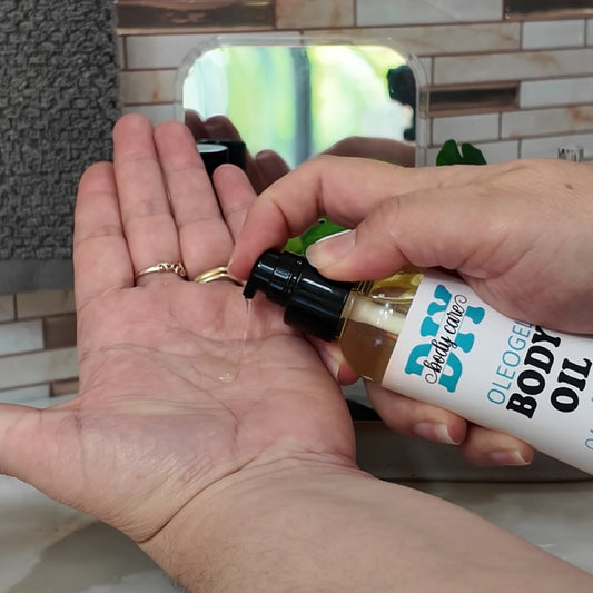 A hand holds a clear bottle with a black serum pump lid. The bottle is filled with a golden liquid which is being pumped out into the palm of another hand. The liquid appears oil like but a little thicker. The bottle is labelled as Oleogel Body Oil.