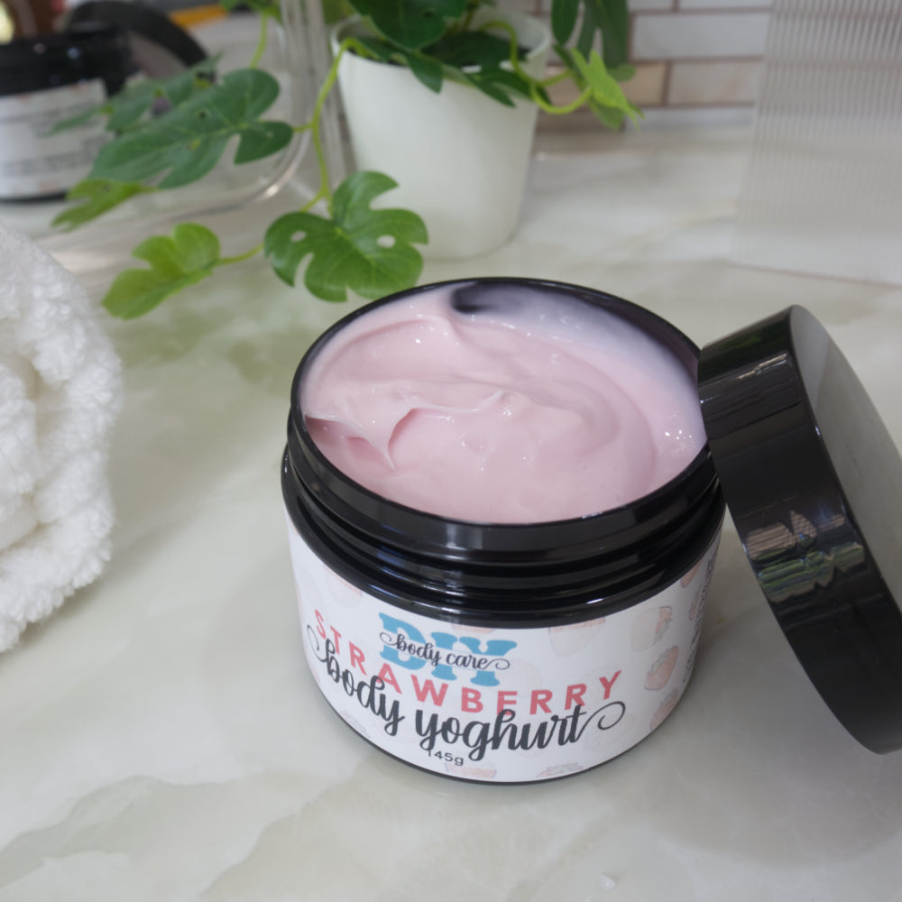 A black jar with a white and pink label sits on a marble counter surounded by a mirror, plant and towel. The black lid has been removed and rests against the side of the jar revealing a pink cream.