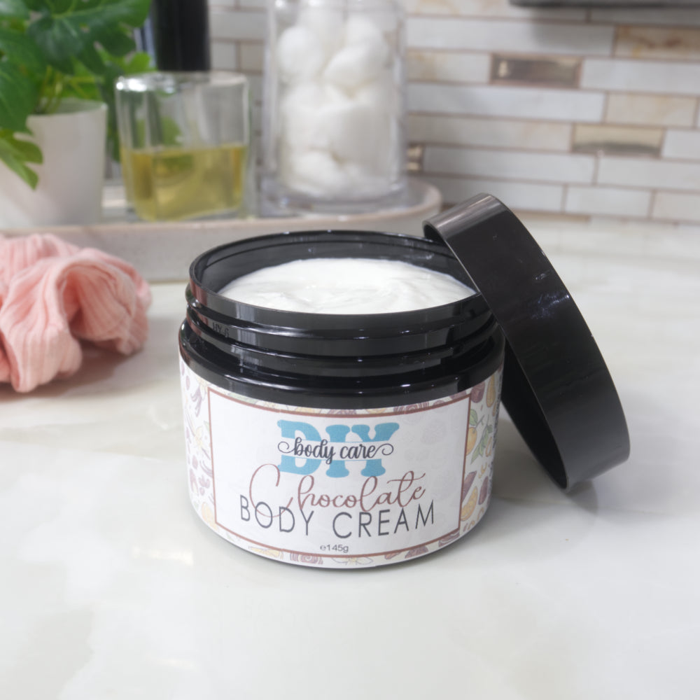 A black jar sits on a bathroom countertop with a blurred background. The jar features a decorative label identifying it as Chocolate Body Cream. The lid has been removed and rests at an angle against the side of the jar revealing a white cream inside