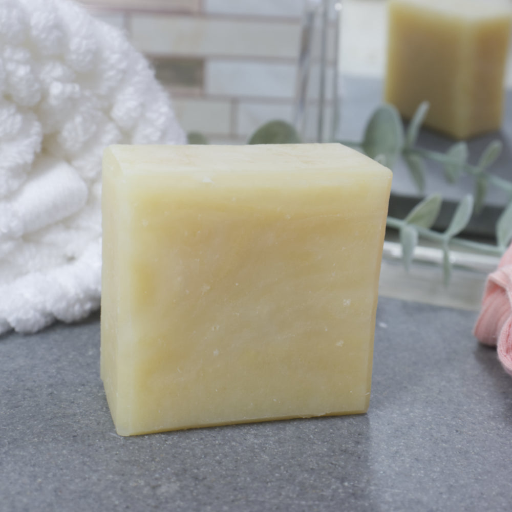 A close up image of a single soap bar. It is square in shape with a pale green colouring. It stands on a grey stone tray surrounded by a white towel and mirror.