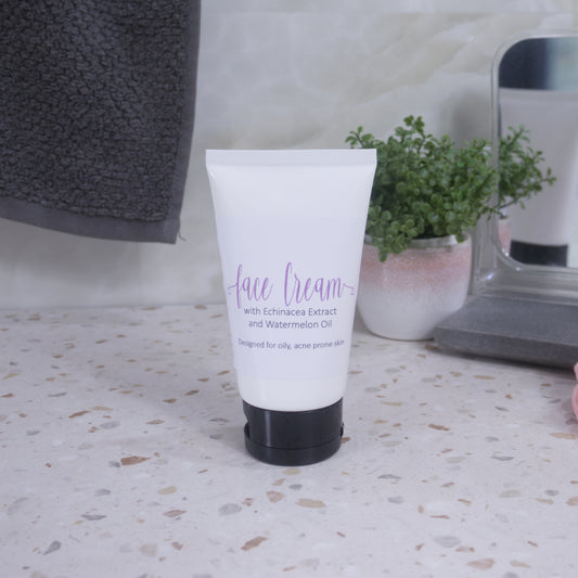 A white tube with black lid and simple product label with text FACE CREAM, stands on a terrazzo bench. Behind is a pottled plant, mirror and a grey towel hanging agains a tiled wall.
