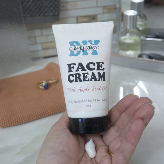 A hand holding a white squeeze tube of DIY body care face cream with apple seed oil, with a small dollop of cream resting below the cap, set against a blurred bathroom countertop background.