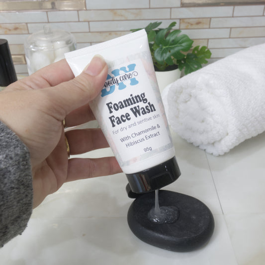A hand holds a white squeeze tube with an open black flip top cap over a black stone on a bathroom countertop. A translucent gel is falling from the tube onto the rock. The tube is listed as Foaming Face Wash.
