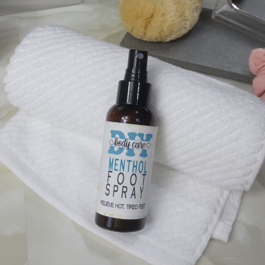 An amber bottle with atomiser nozzle and white product label lies resting against a rolled up towel on a marble surface. Glimpses of a body brush, mirror and scrunchie can be seen in the background.