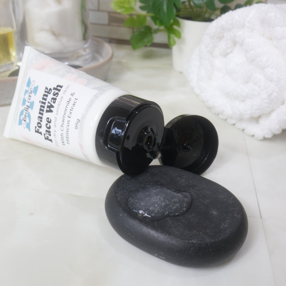 To the front is a black flat stone with a small pool of translucent, gel like liquid. Resting on the edge of stone is a white tube with a black flip top cap that is open. The tube is labeled as foaming face wash.
