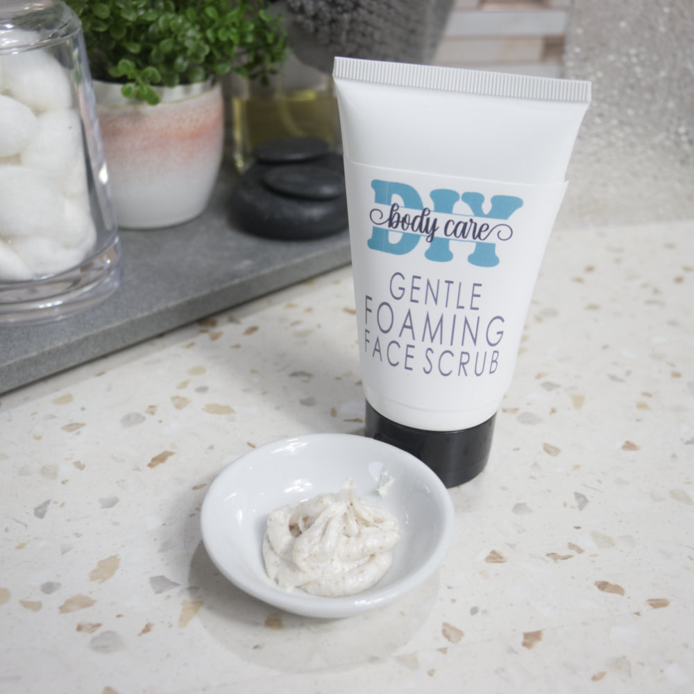 A white tube with black flip-top cap stands on a terrazzo bathroom countertop next to a stone tray with a plant, cotton balls and perfume bottle. The tube is labelled as GENTLE FOAMING FACE SCRUB. To the front sits a small with dish with some of the scrub in it. The scrub is an off white colour with flecks of brown botanicals.
