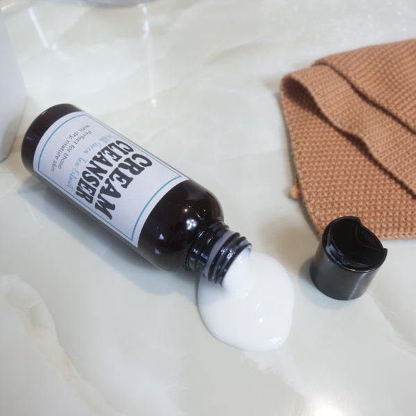 An amber bottles lies flat on a marble tie surface with the disc cap sitting to the side next to an orange cloth. A white lotion flows out of the bottle onto the countertop.