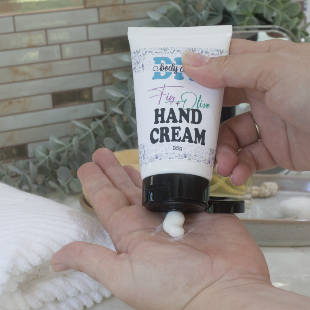 A hand squeezes some white cream into the palm of another hand from a white tube with black flip top lid. The tube has a product label identifying it has Hand Cream. The background showings a white tile and greenery