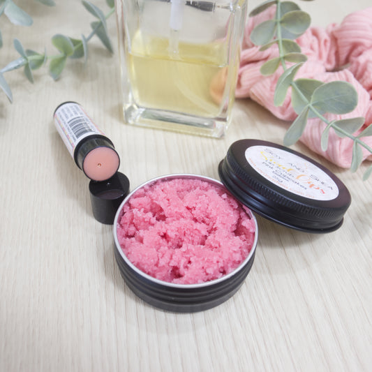 A round black tin sits on a wooden surface next to a black tube of lip balm. In the background is a perfum bottle, scrunchie and greenery. The lid of the tin rests on the side of the tin with label reading SWEET LIPS LIP SCRUB. The tin is filled with a pink sugar scrub. The Lip balm has had its cap removed and the tube is resting on the cap revealing a pink balm