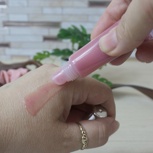A hand squeezes a dusky pink lip oil from a small tube onto the back of the other. The oil on the hand has a slight pink hue with the skin still visable under the oil. The background is blurred with tiles and a plant