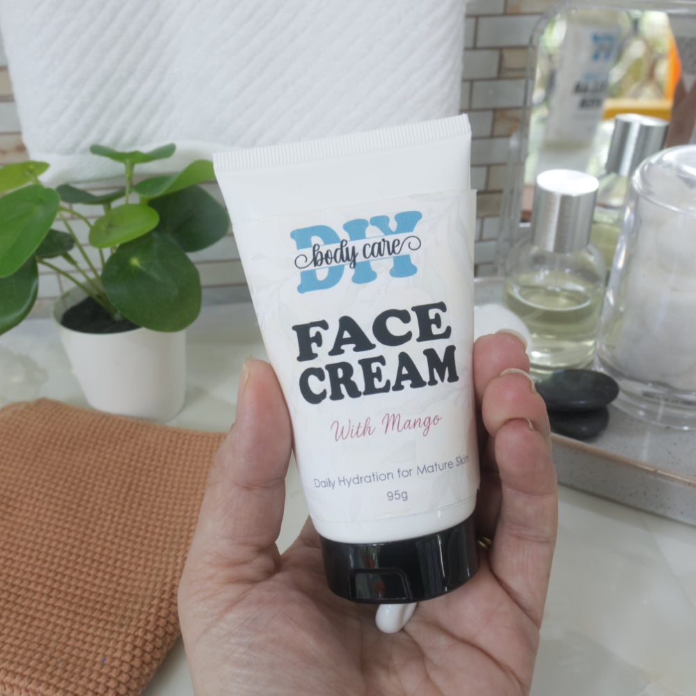 A hand holding a white squeeze tube of DIY body care Face Cream with Mango, set against a bathroom counter background with a small potted plant and skincare bottles.