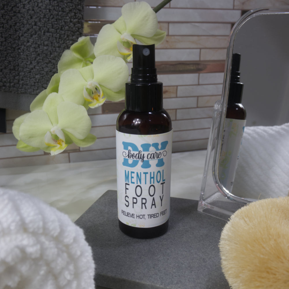 An amber bottle with atomiser sprayer and white product label stands on grey stone tray next to mirror and a spray of yellow orchids. In the background is a towel hanging against a tiled wall.