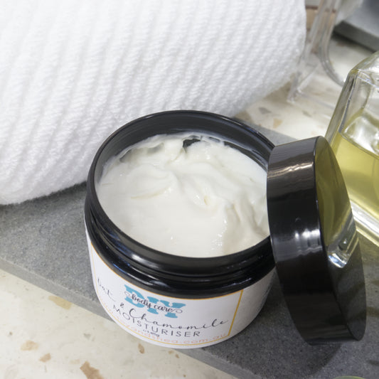 A black jar with the lid resting on the side revealing an off white cream stands on a grey stone tray next to a white towel and bottle of perfume.