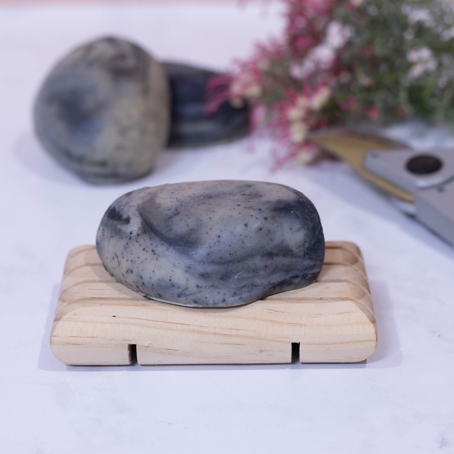 A soap in the shape of a stone with black and white marblings sits on a wooden soap rack. Behind, in a blurred backgroudn are two other soaps plus a bunch of flowers and secateurs