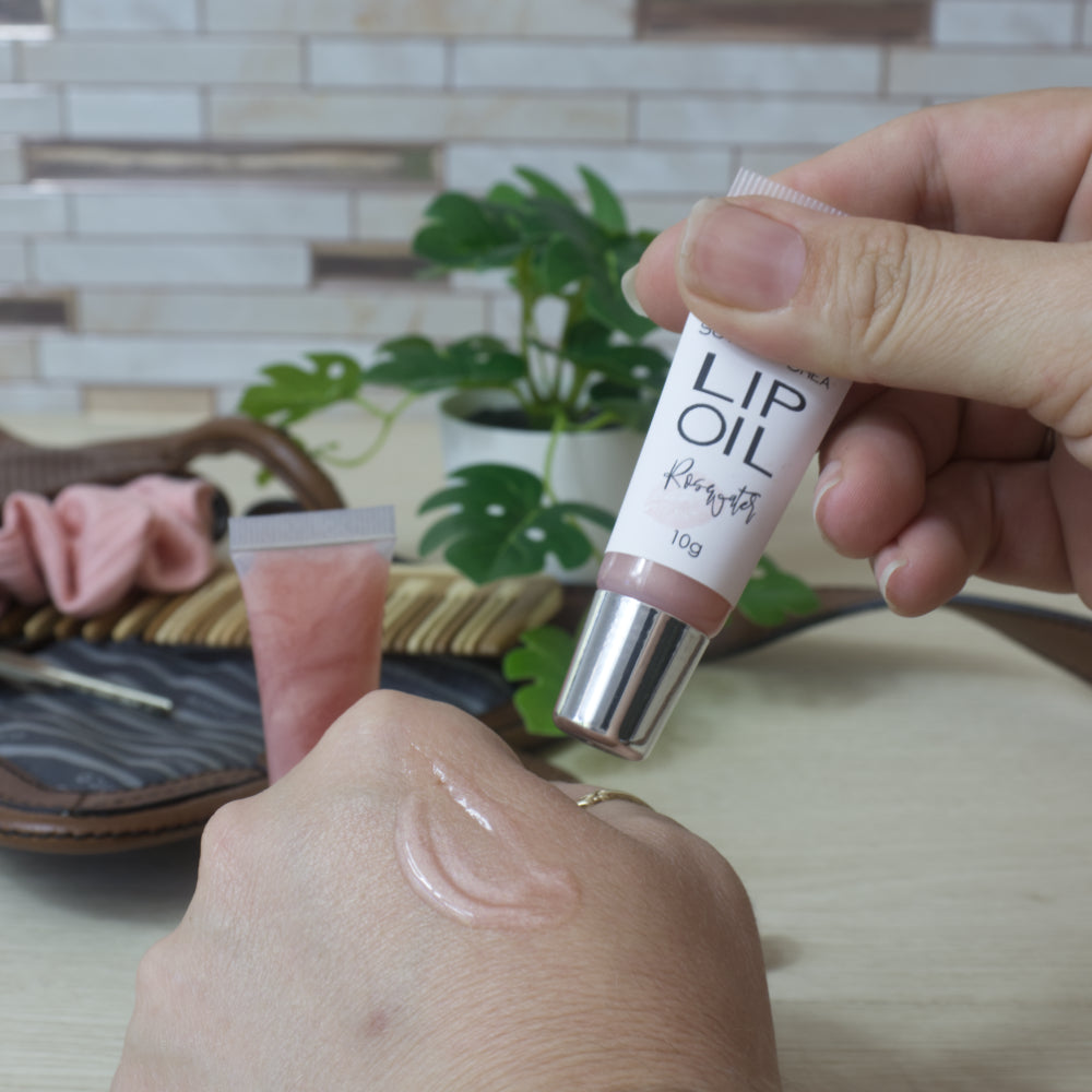 A hand holds a tube labelled Lip OIl above the back of another hand which has some of the shiny lip oil on it. In the background is counter top with a small handbag and potted plant.