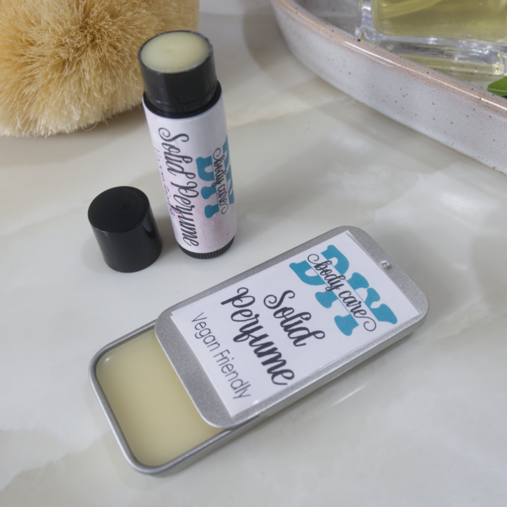 Two solid perfume are sitting on a bathroom countertop. One is presented in a rectangular silver tin with a open sliding lid while the other is a black tube like lip balm, standing upright with the lid to the side of the tube revealing white balm inside.