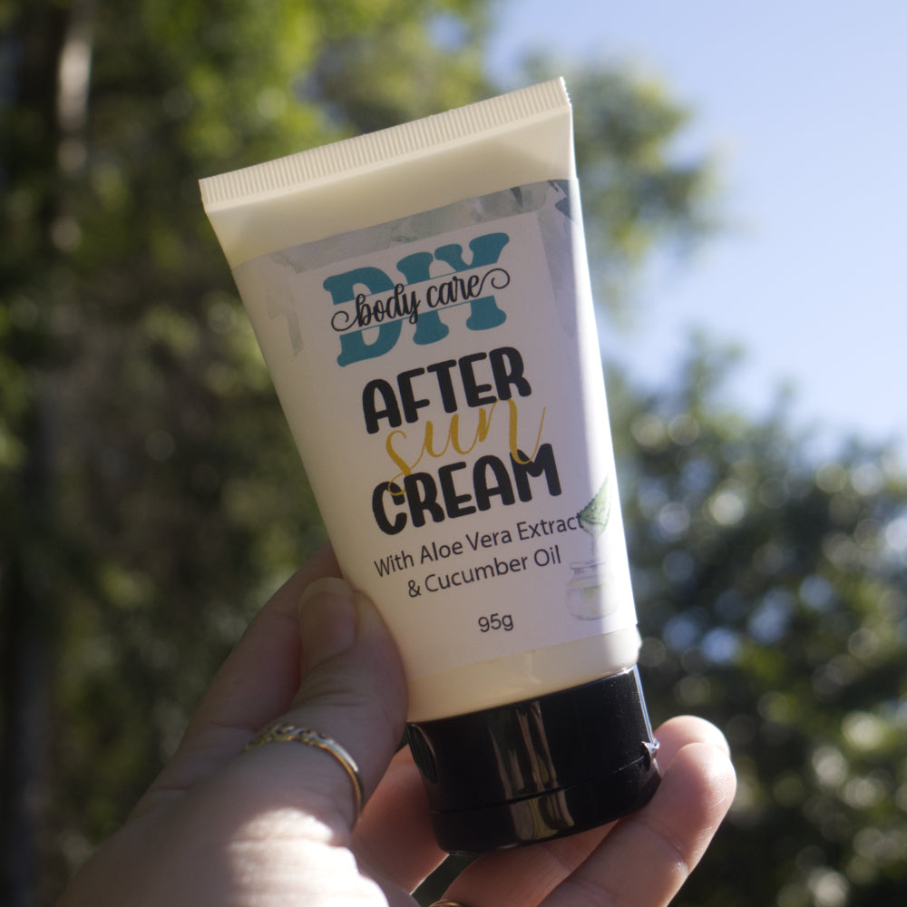 A hand holds a a white tube with black lid up in the air with a blurred background of trees and blue sky with the sun rays shining on the tube.  The tube features a product label reading AFTER SUN CREAM