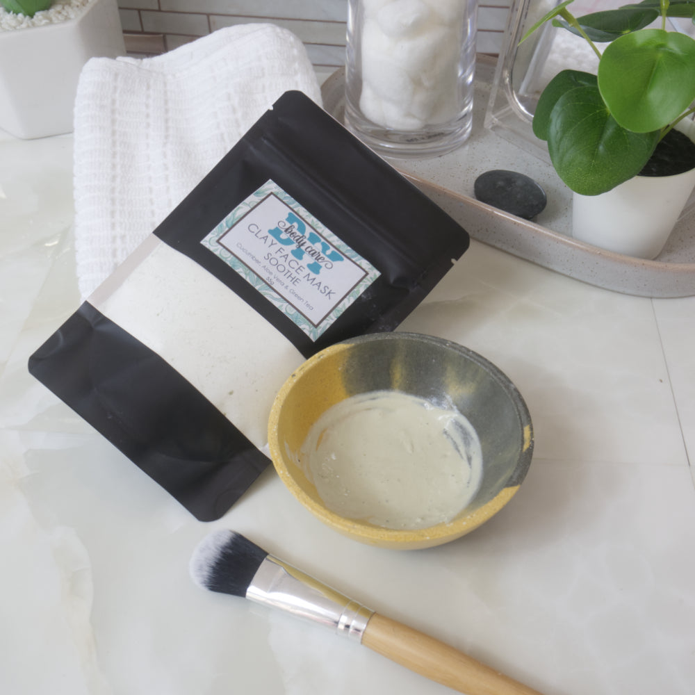 A black stand up pouch with clear window lies resting against a white towel.  Through the window, a cream coloured powder can be seen.  To the right is a small bowl which has a pale green paste in it as well as a facial brush