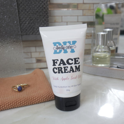 A white squeeze tube of DIY body care Face Cream with Apple Seed Oil and a black flip-top cap, standing on a bathroom counter with a mirror, soap dish, and small bottles in the background.