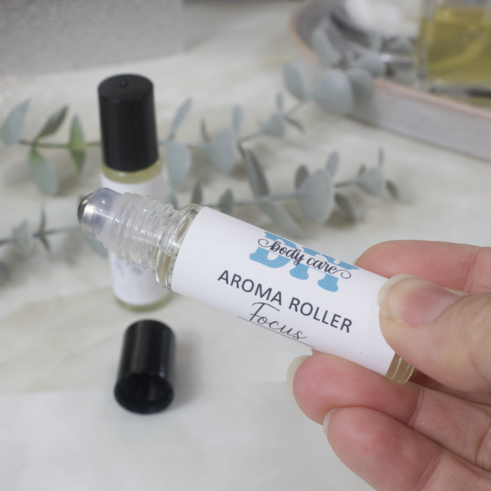A hand holds a clear glass aromatherapy roller bottle with a white label reading 'DIY body care' and 'AROMA ROLLER Focus'. Another bottle with a black cap sits in the background on a light surface with eucalyptus leaves.