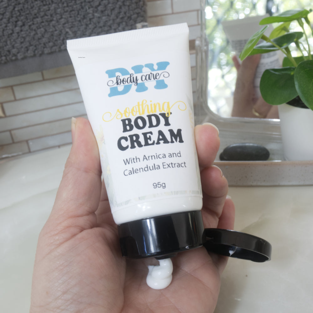 A hand holding a white squeeze tube of DIY body care soothing body cream with an open black cap, with a small amount of cream squeezed out. The background shows a bathroom counter and a plant.