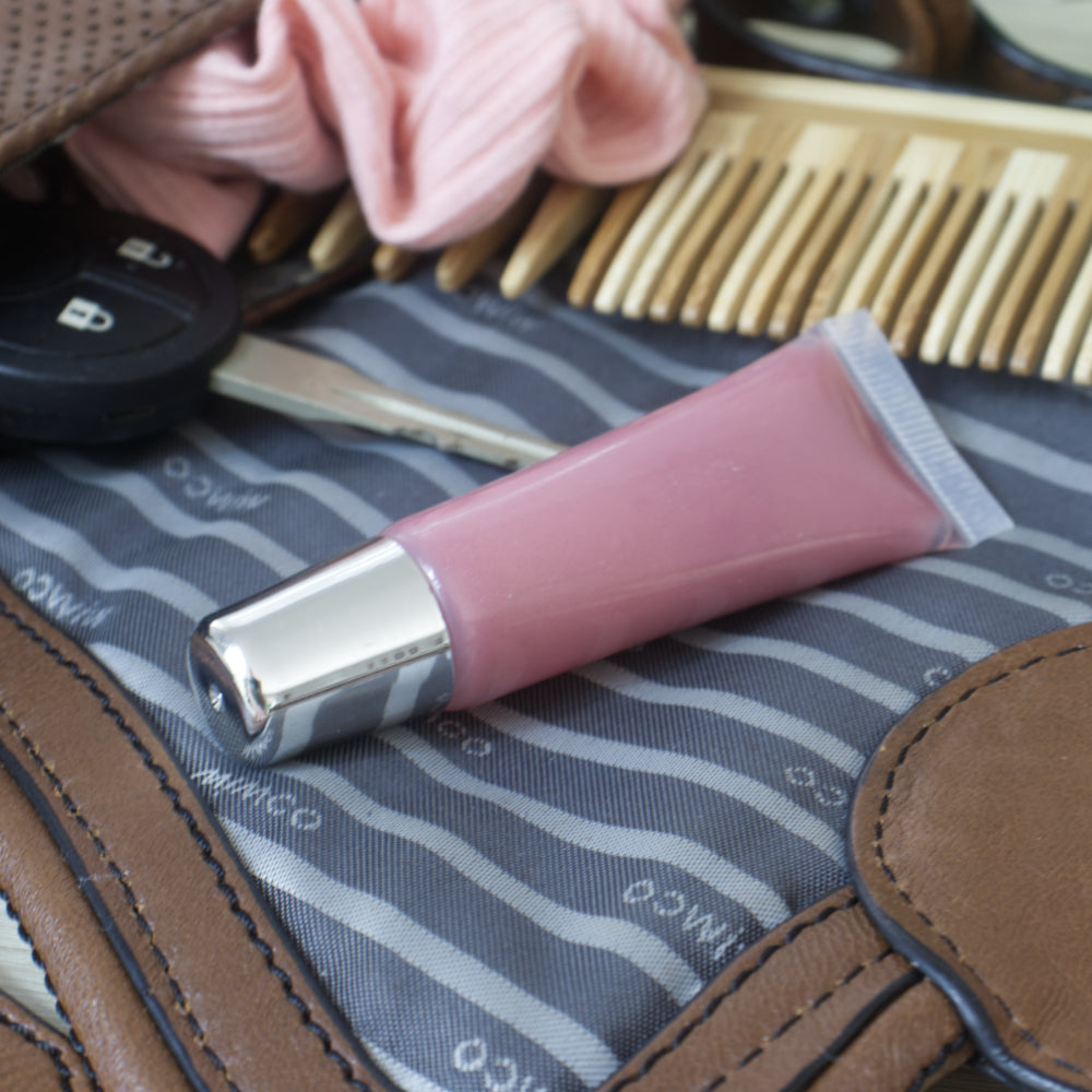 A clear tube with a silver lid is filled with a dusky pink lip oil.  It lies on the flap of a handbag that lies open with its content flowing out.