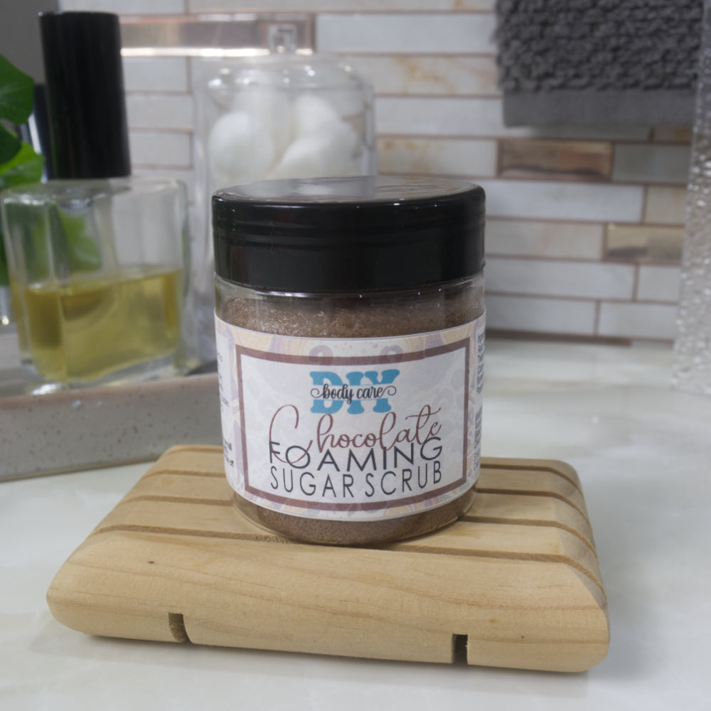 A clear jar with black lid sits on a wooden soap rack on a bathroom countertop.  It is labelled a chocolate Foaming Sugar Scrub.  The base of the jar is clear revealing a chocolate coloured scrub inside