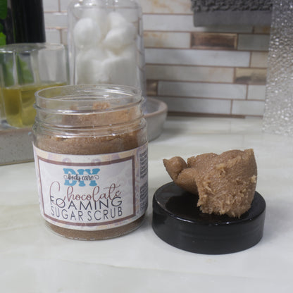 A clear jar labelled as Chocolate Foaming Scrub sits on a bathroom countertop.  The black lid has been removed and sits beside the jar with a piece of the chocolate scrub sitting on top of it