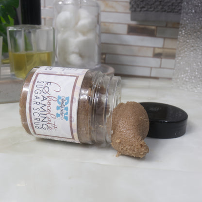 A clear jar labelled as Chocolate Foaming sugar scrubs lays on its side on a bathroom counter top.  The black lid has been removed and placed beside it and a piece of the scrub has fallen out of the jar.  The scrub has a chocolate colour