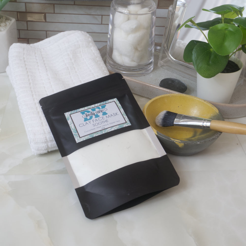 A black standup pouch with a clear window pane lays resting against a white towel on a bathroom countertop.  The pouch is labelled as Clay Face Mask Sooth and the window shows a very pale green powder inside.  Next to the pouch is a small yellow and grey bowl with a facial brush