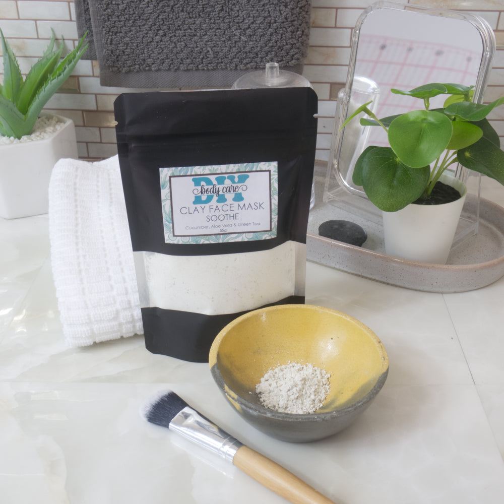 A black standup pouch with clear window is standing upright on a bathroom counter top and is labelled as Clay Face Mask.  Behind the pouch is a white towel and in front is a small yellow and grey bowl that has some dry powder in it.  The powder is a very pale green