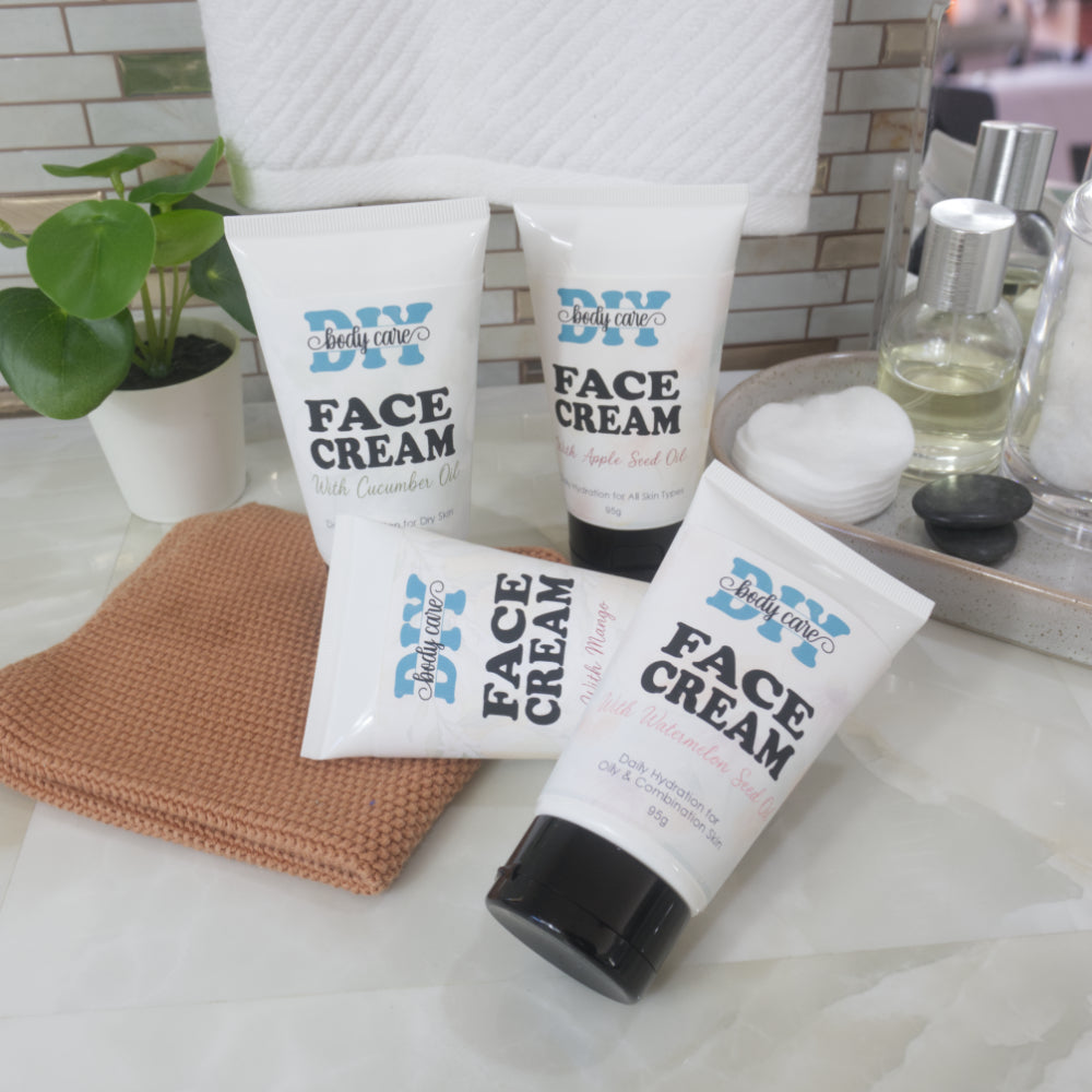 Four white tubes with black caps are presented on a bathroom countertop surrounded by accessories and a plant.  Each tube is labelled as Face Cream but each one has a different fruit oil listed on it along with the skin type it is suited for