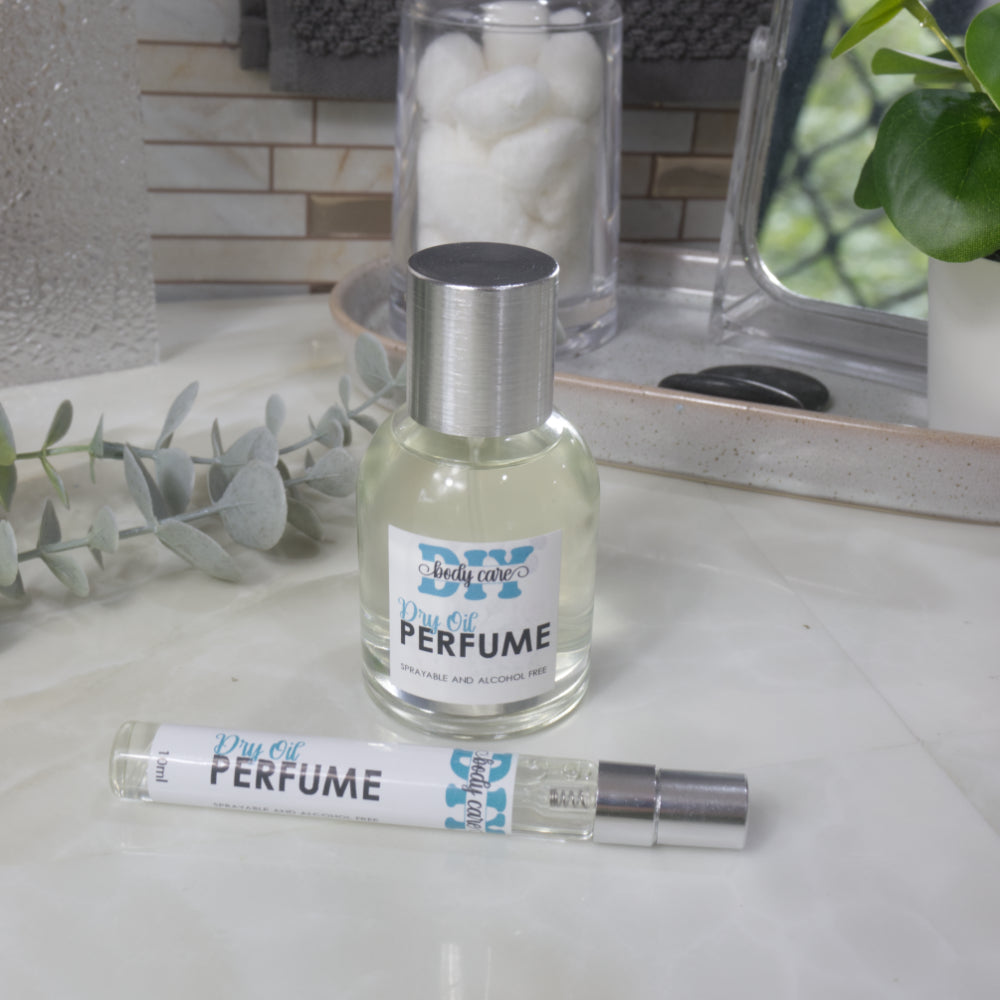 A small clear round bottle of perfume with a silver cap stands on a light marble surface.  Placed in front is a long narrow 10ml spray bottle. The label reads 'DIY Body Care Dry Oil Perfume'.