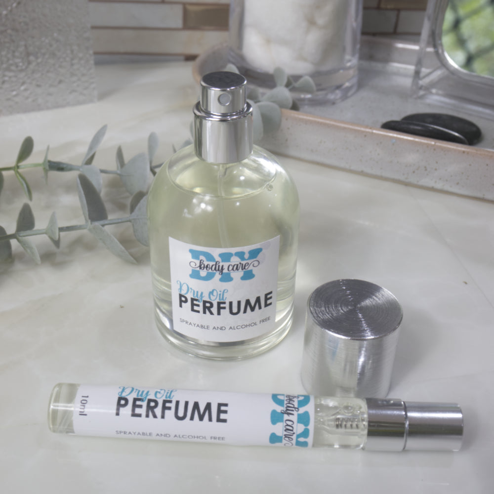 A round clear bottle of Dry Perfume Oils stands on a bathroom countertop with the silver lid removed and placed to the side revealing a silver spray atomiser.   In front lays a long 10ml bottle of perfume with silver cap