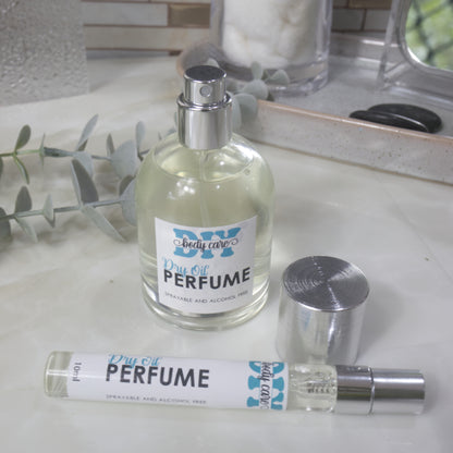A round clear bottle of Dry Perfume Oils stands on a bathroom countertop with the silver lid removed and placed to the side revealing a silver spray atomiser.   In front lays a long 10ml bottle of perfume with silver cap