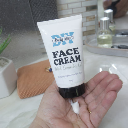 A hand holding a white squeeze tube of DIY body care face cream with cucumber oil, with the white cream being squeezed into the palm of the hand.  Behind is a a marble surface in a bathroom setting.