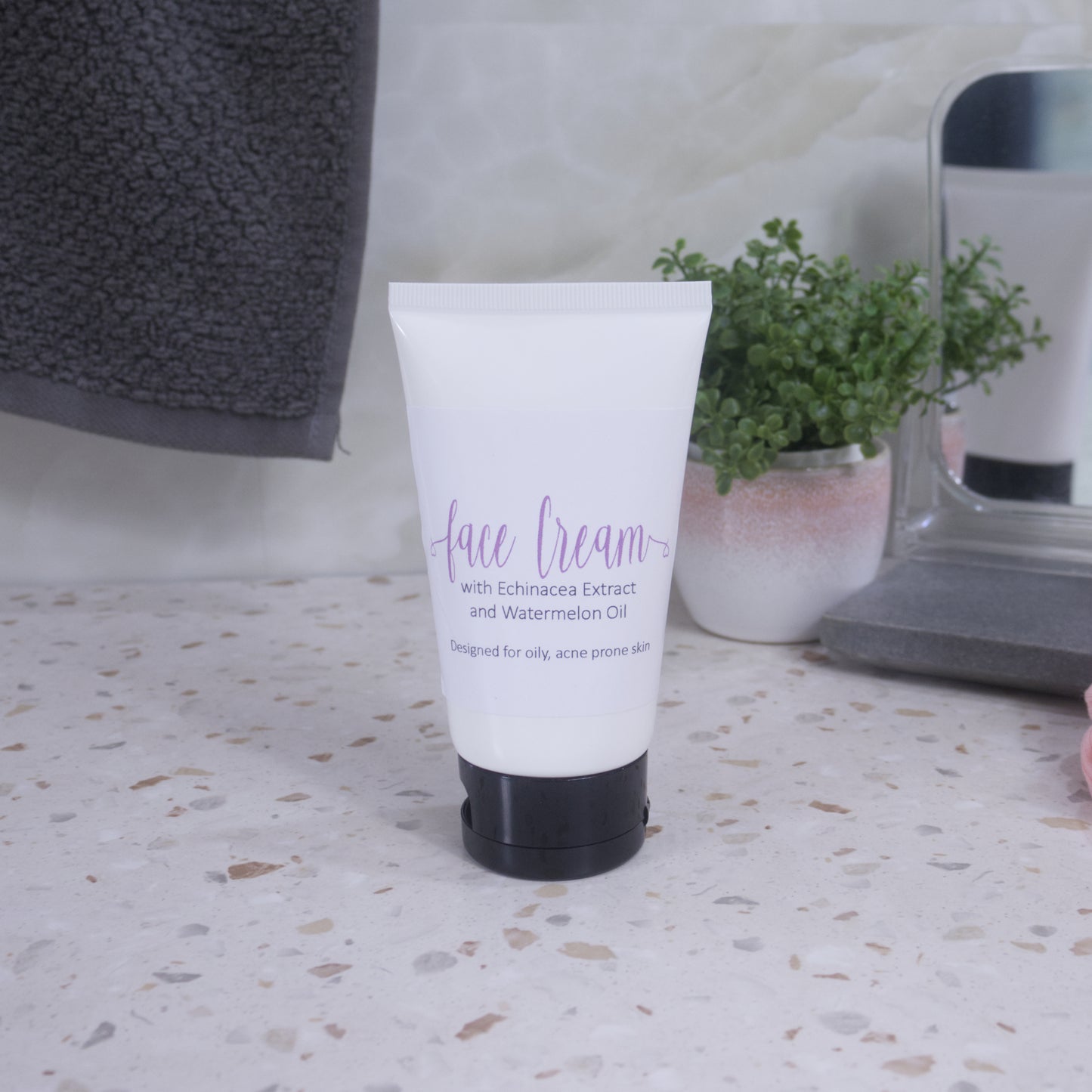 A white tube with black lid and simple product label with text FACE CREAM, stands on a terrazzo bench. Behind is a pottled plant, mirror and a grey towel hanging agains a tiled wall.