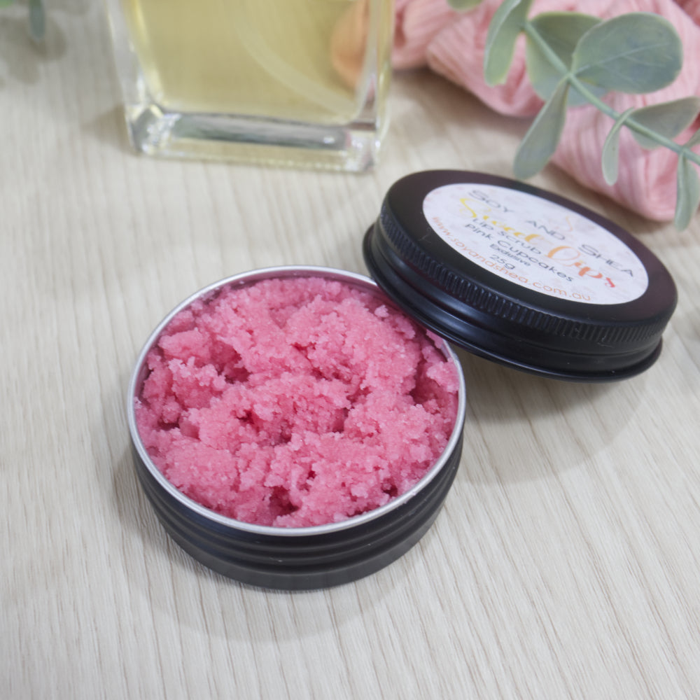 A round black tin sits on a wooden surface with a bottle of liquid and pink cloth in the background.  The lid of the tin rests on the side of the tin with label reading SWEET LIPS LIP SCRUB.  The tin is filled with a pink sugar scrub.