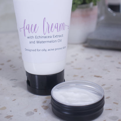 A close up image of a white tube with black lid labelled FACE CREAM sitting on a terrazzo countertop with a blurred bathroom background.  In the foreground the image is focused ona small black tin filled with white cream.
