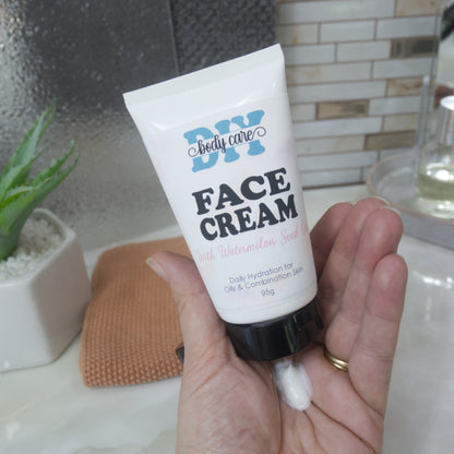 A hand holding a white squeeze tube of DIY body care face cream with a black cap, with a small dollop of white cream dripping from the bottom, set against a bathroom background with a small plant and a textured towel.