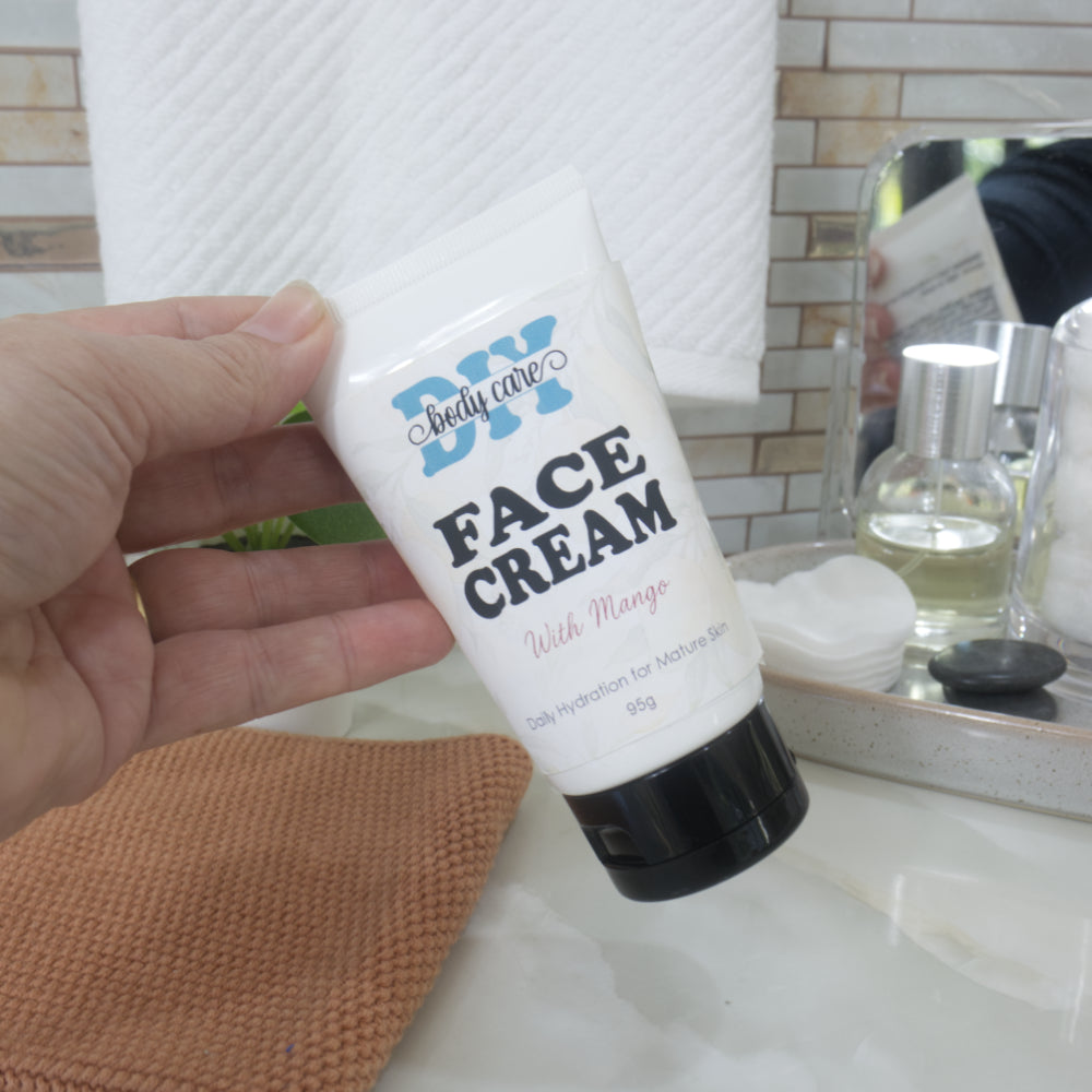  A hand reaches in from the left to pick up a white tube with black cap up from a bathroom counter top.  The tube is labelled as Face Cream with Mango, for Mature skin