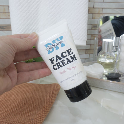  A hand reaches in from the left to pick up a white tube with black cap up from a bathroom counter top.  The tube is labelled as Face Cream with Mango, for Mature skin