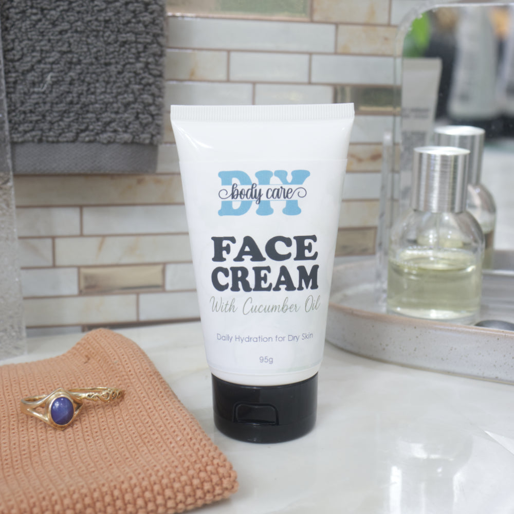 A white squeeze tube of DIY body care Face Cream with cucumber oil, featuring a black cap, standing on a white marble countertop. The background shows a tiled bathroom wall, a peach-colored towel with a gold ring, and a mirror reflecting a glass bottle.