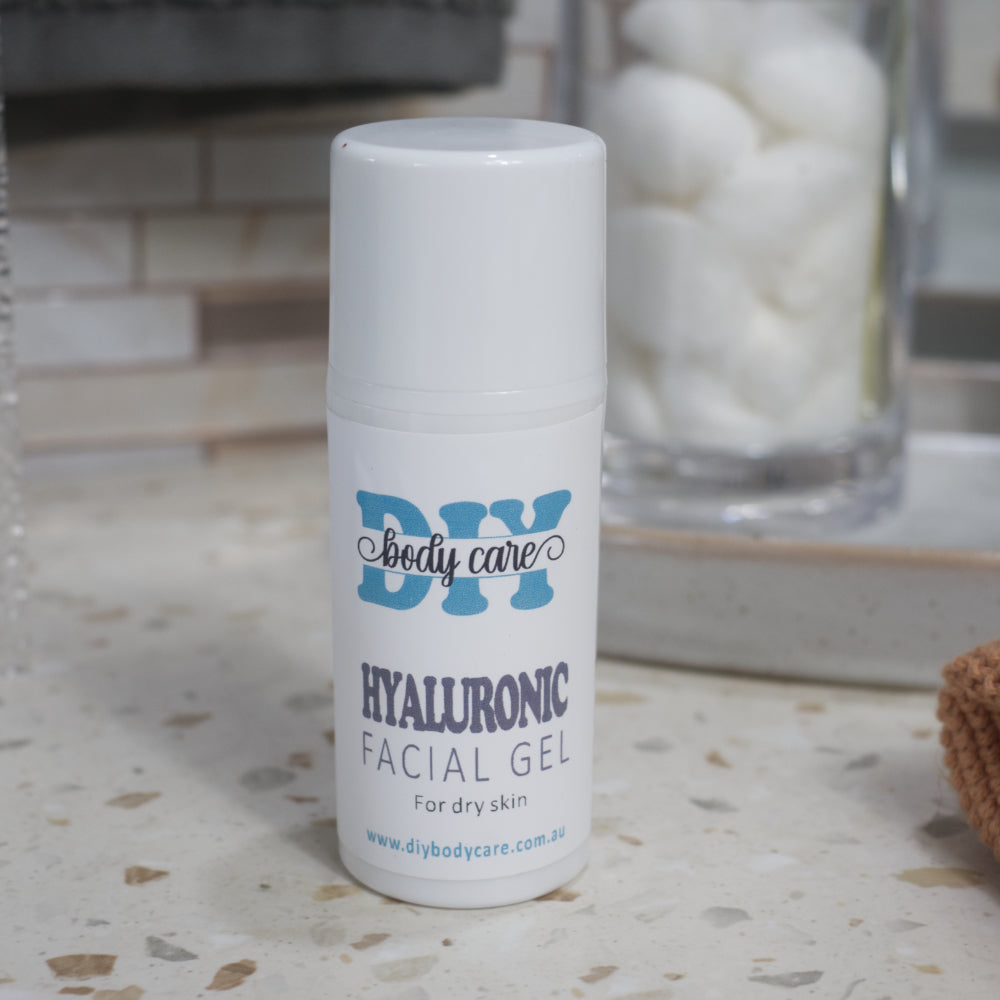 A white bottle with cap labelled DIY Body Care Hyaluronic Facial Gel, sits on a terrazzo countertop with a blurred background where a jar of cotton balls can be made out.