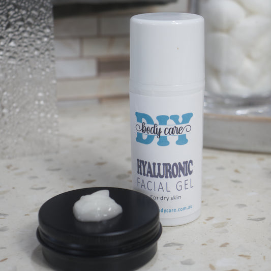 A white bottle with cap stands on a terrazzo bench top with a product label reading HYALURONIC FACIAL GEL.  Behind sis a blurred background wth cotton balls.  In the foreground is a small black tin with some Facial Gel on it.  The gel is a white.