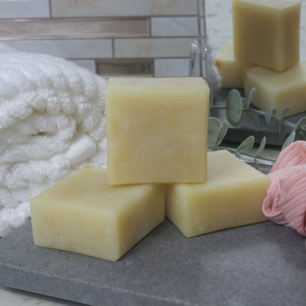 Two pale green square soaps lay flat on a grey stone tray with a third soap standing upright upon them.   In the background is a white towel and a small mirror reflecting the soaps