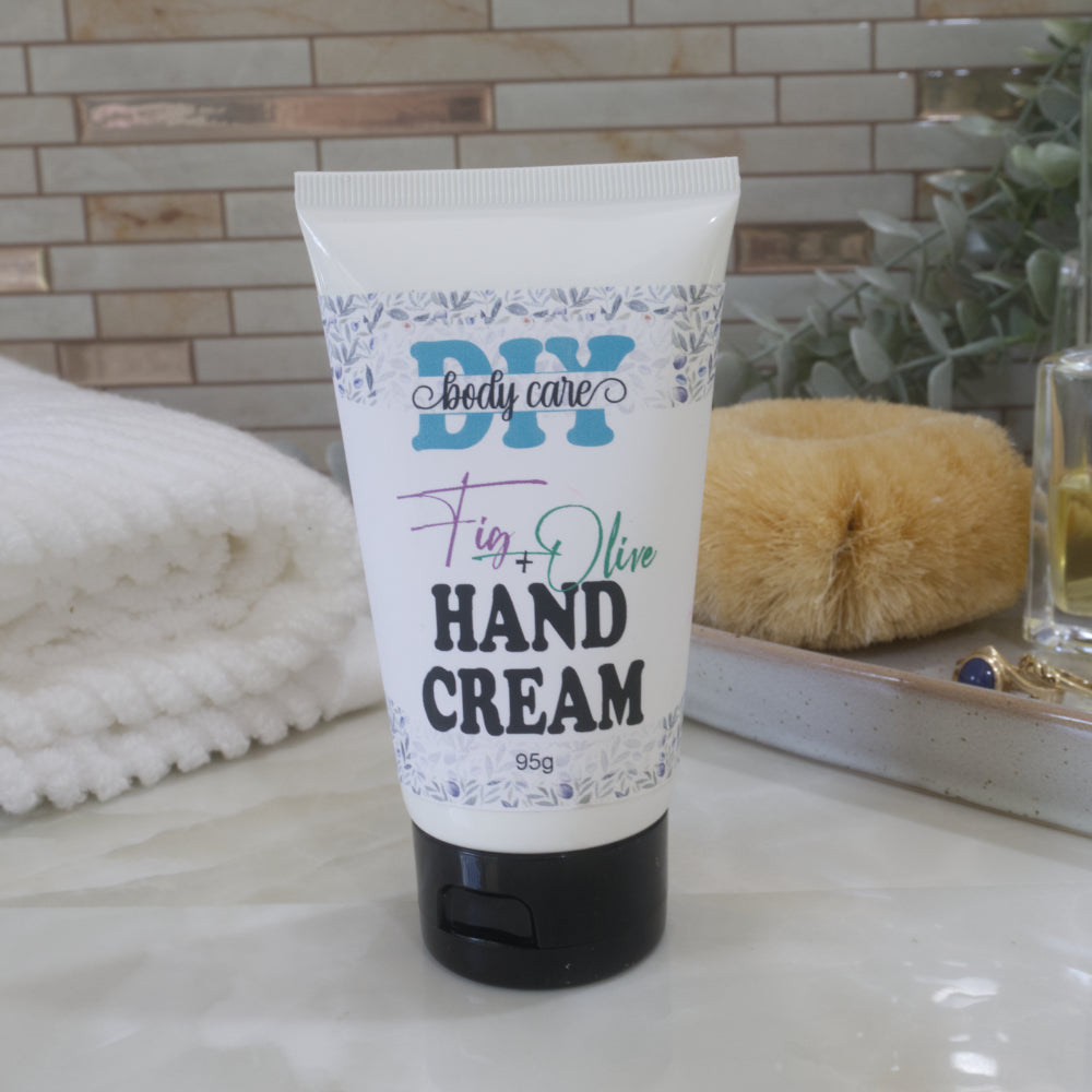 A white tube with black flip top cap sits on a marble bathroom counter top with a white towel, body brush and greenery against a tiled wall.  The tube features a product label identifying it as Hand Cream