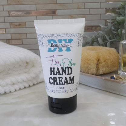 A white tube with black flip top cap sits on a marble bathroom counter top with a white towel, body brush and greenery against a tiled wall.  The tube features a product label identifying it as Hand Cream